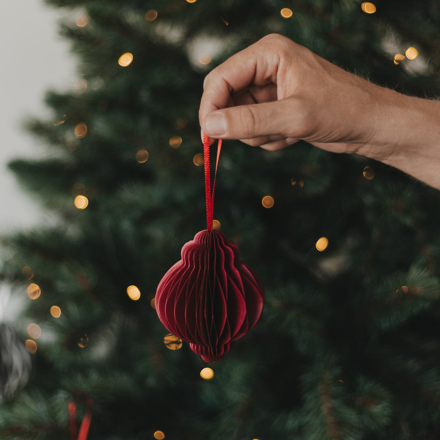 Roter Papieranhänger in Kugelform wird vor einem beleuchteten Weihnachtsbaum gehalten