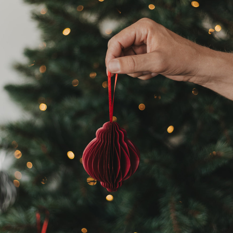 Roter Papieranhänger in Kugelform wird vor einem beleuchteten Weihnachtsbaum gehalten