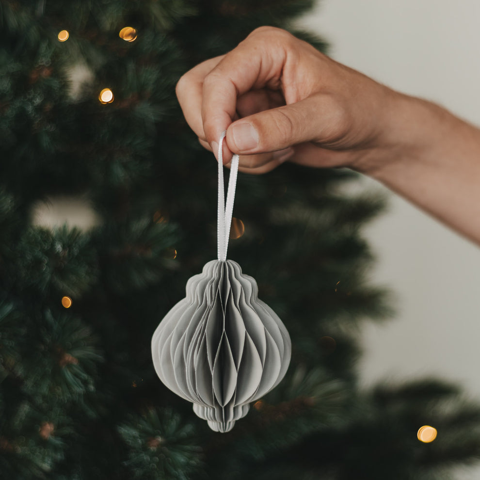Grauer Papieranhänger in Kugelform wird vor einem beleuchteten Weihnachtsbaum gehalten