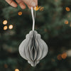 Grauer Papieranhänger in Kugelform wird vor einem beleuchteten Weihnachtsbaum präsentiert