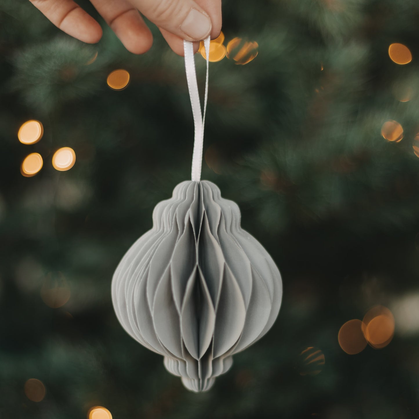 Grauer Papieranhänger in Kugelform wird vor einem beleuchteten Weihnachtsbaum präsentiert