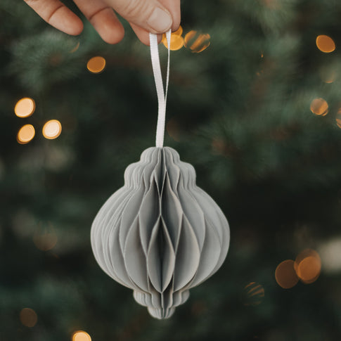 Grauer Papieranhänger in Kugelform wird vor einem beleuchteten Weihnachtsbaum präsentiert