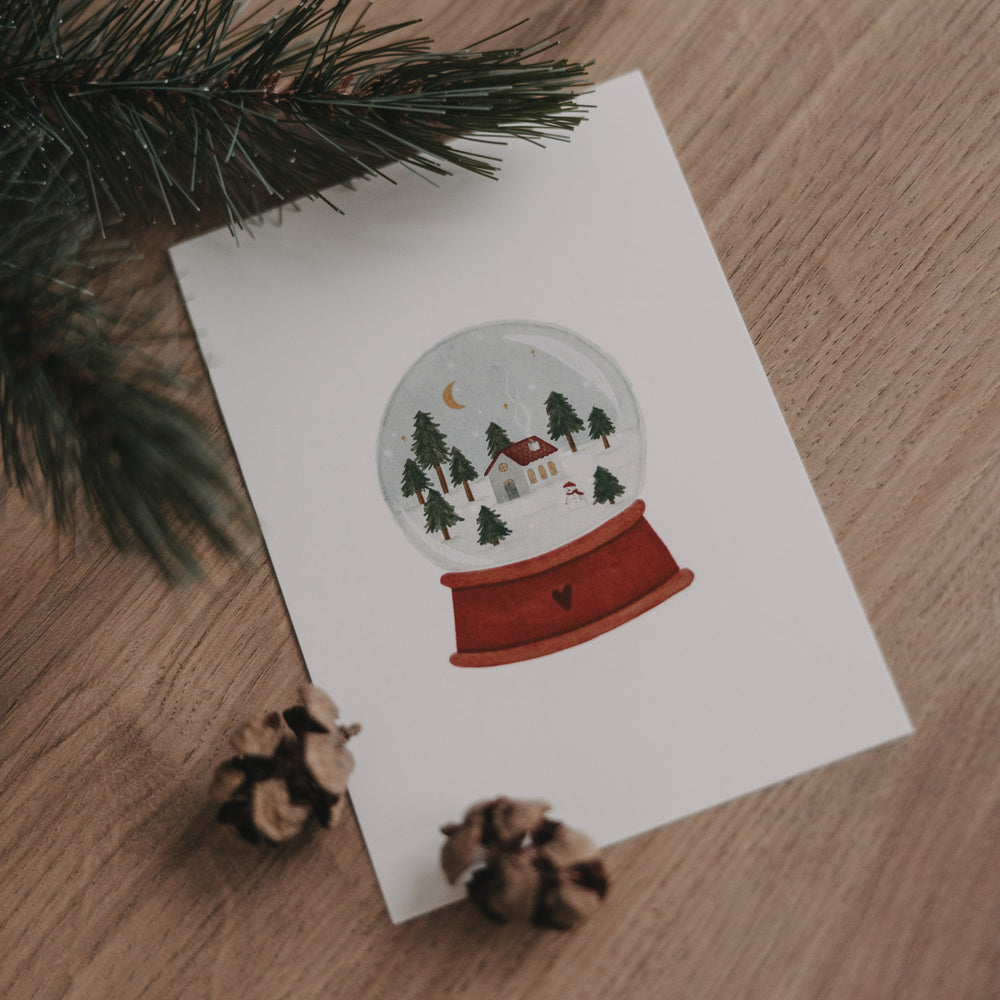 Weihnachtskarte mit Schneekugel, Häuschen, Bäumen und Mond auf rotem Sockel