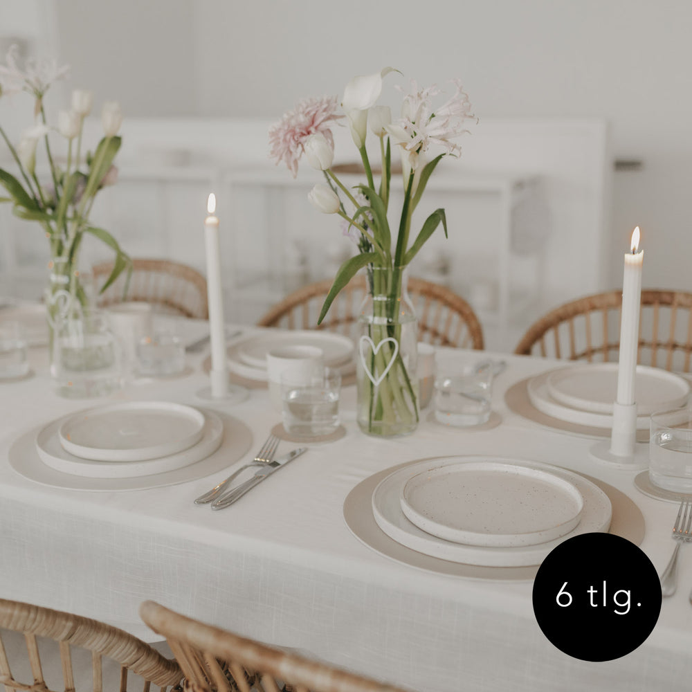 Hell gedeckter Tisch mit cremefarbenen Platzsets, Keramikgeschirr und frischen Blumen in Glasvasen.