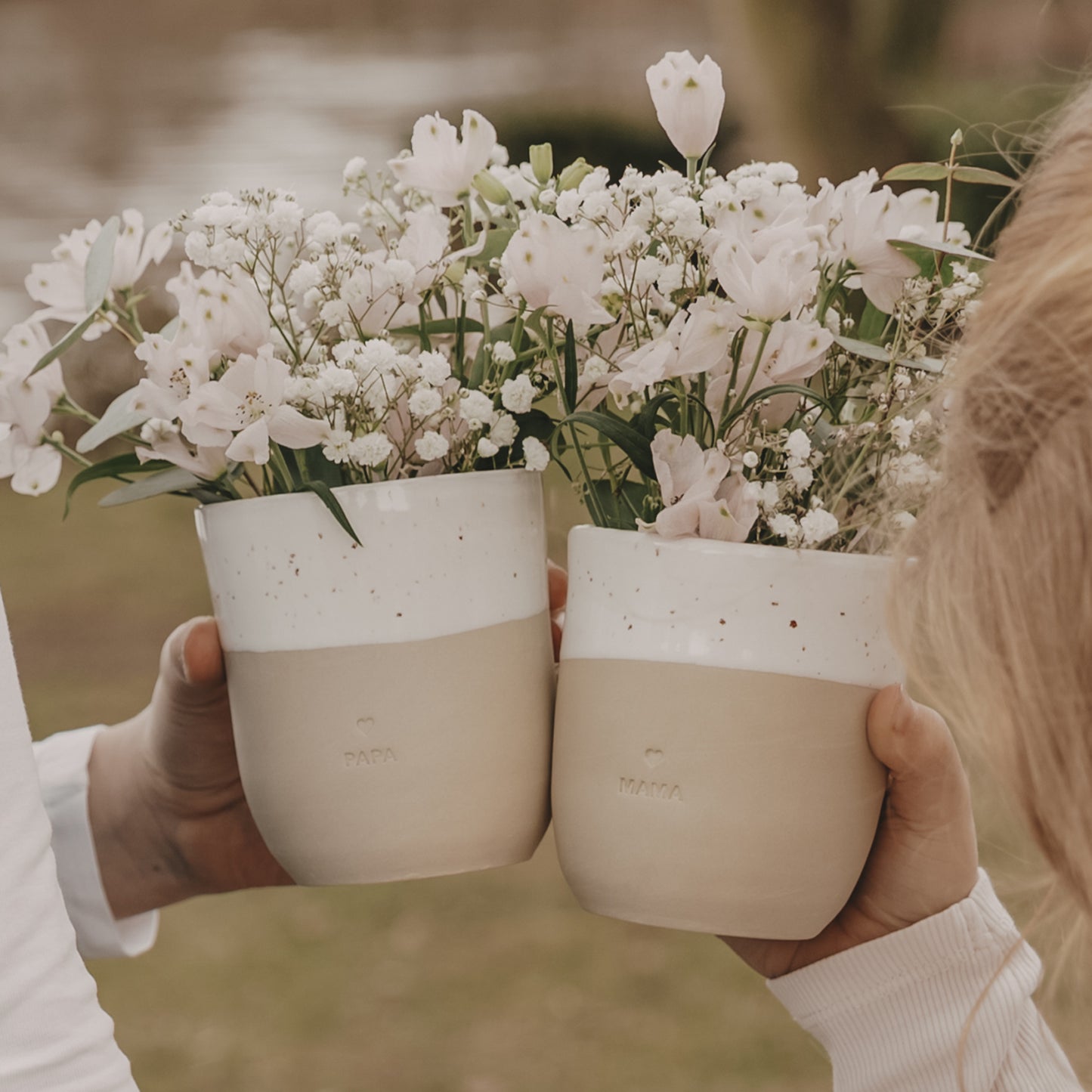 Ein Becher mit der Prägung "Mama" wird von einem süßen Mädchen gehalten und stößt mit einem Becher mit der Prägung "Papa" an.