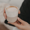 Nahaufnahme eines zweifarbigen Keramikbechers „Me Time“ mit Herz und Cappuccino, der in beiden Händen gehalten wird
