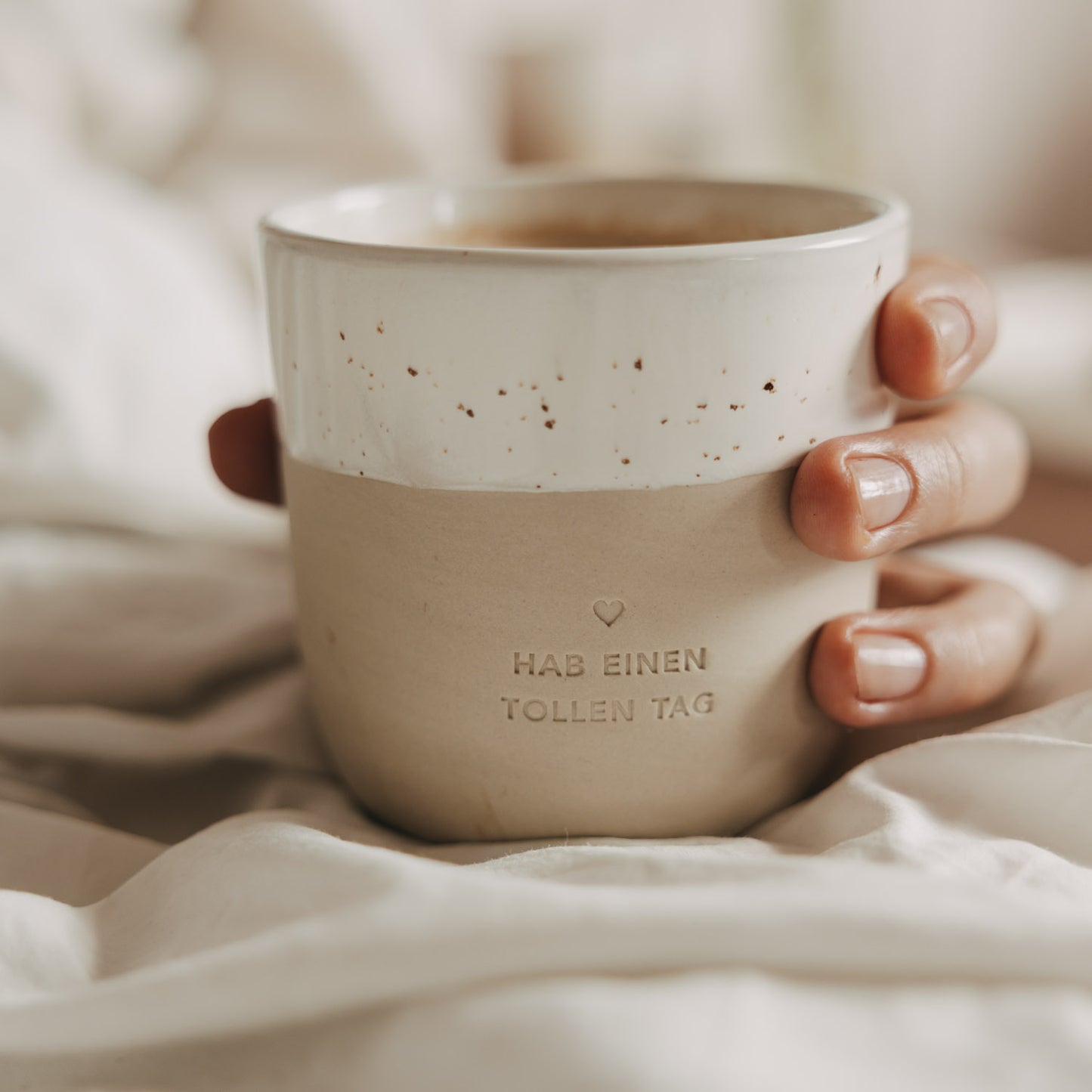 Keramikbecher „Hab einen tollen Tag“ in Beige und Weiß mit Kaffee, gemütlich im Bett gehalten