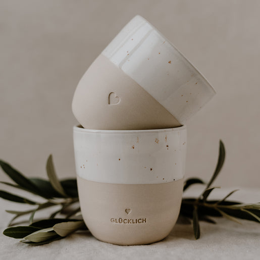 Zwei Becher aus Keramik, einer leicht in den anderen gelehnt. Vorderer Becher trägt die Prägung ‚GLÜCKLICH‘. Dekorative Olivenzweige ergänzen das natürliche Setting.