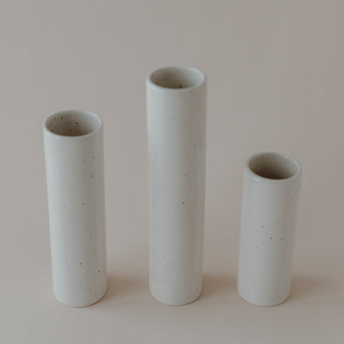 Drei zylindrische, minimalistische Keramikvasen in verschiedenen Höhen stehen nebeneinander auf einem hellen, neutralen Untergrund. Die cremeweißen Vasen mit feinen Sprenkeln wirken schlicht und modern, ohne Blumen oder Dekoration.