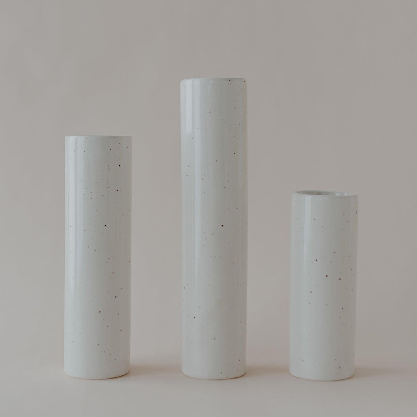 Drei schlichte, zylindrische Keramikvasen in verschiedenen Höhen stehen nebeneinander auf hellem Untergrund. Die Vasen sind cremefarben mit feinen, unregelmäßigen Sprenkeln und haben eine glänzende Oberfläche.