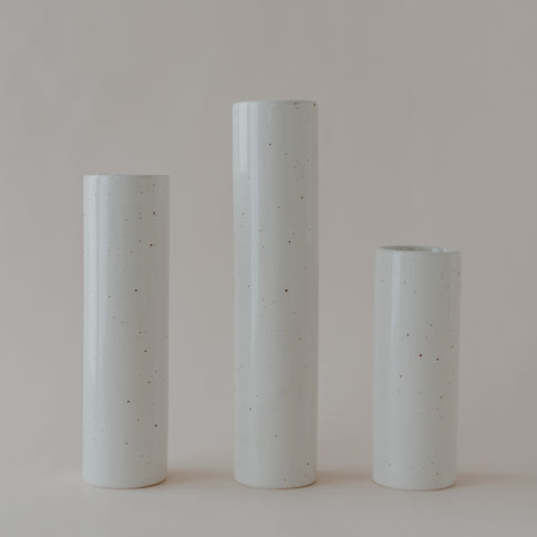 Drei schlichte, zylindrische Keramikvasen in verschiedenen Höhen stehen nebeneinander auf hellem Untergrund. Die Vasen sind cremefarben mit feinen, unregelmäßigen Sprenkeln und haben eine glänzende Oberfläche.
