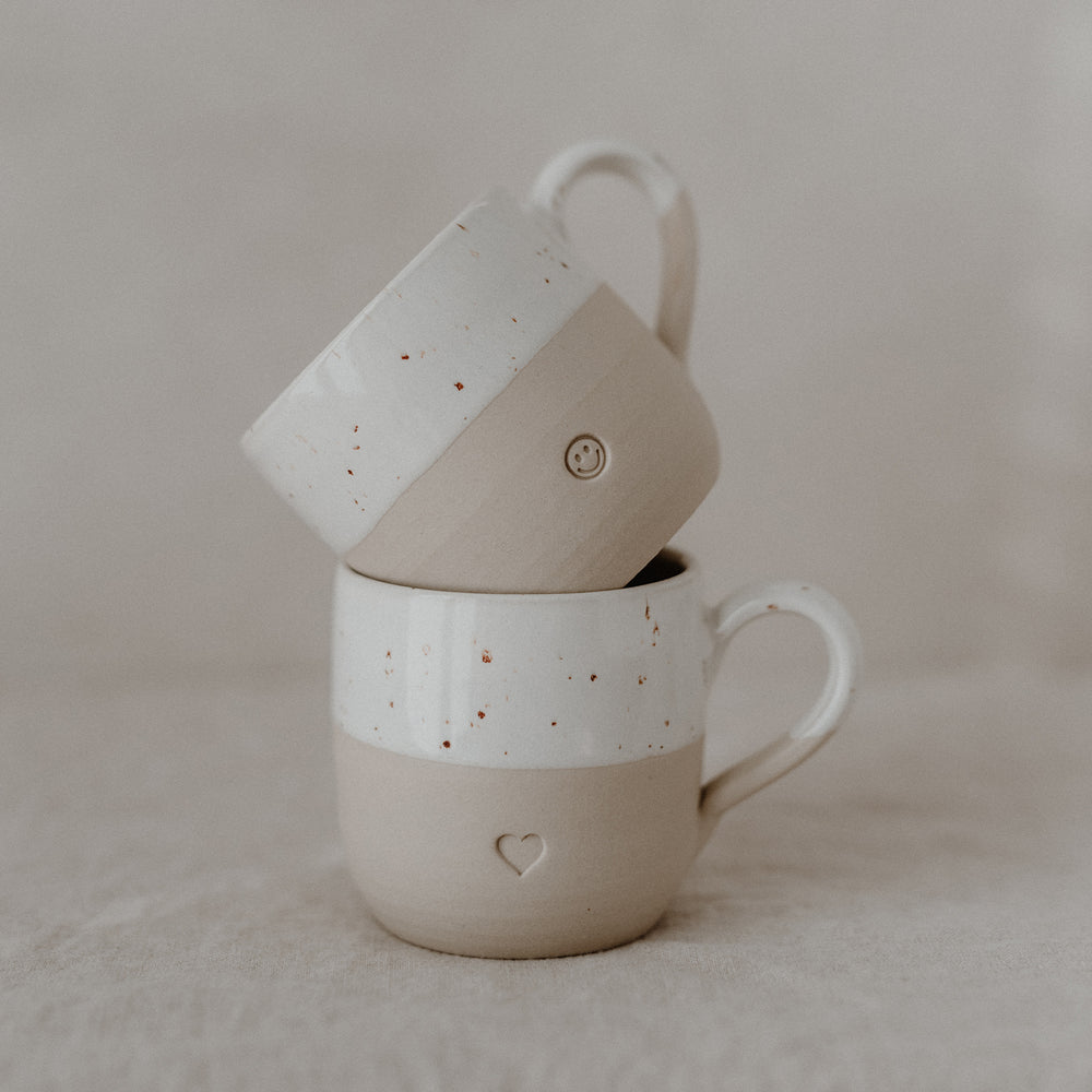 Zwei beige-weiße Keramiktassen mit gesprenkelter Glasur sind aufeinander gestapelt und stehen auf hellem Stoff. Die untere Tasse trägt eine eingeprägte Herzform, die obere ein kleines Smiley-Gesicht. Beide Tassen haben Henkel und zeigen eine liebevolle, minimalistische Gestaltung – harmonisch präsentiert mit sanften Naturtönen.