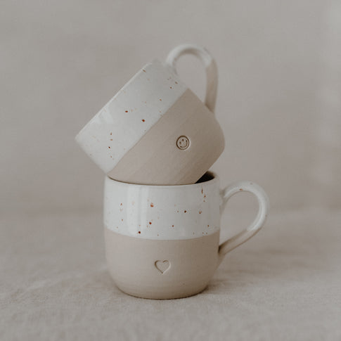 Zwei beige-weiße Keramiktassen mit gesprenkelter Glasur sind aufeinander gestapelt und stehen auf hellem Stoff. Die untere Tasse trägt eine eingeprägte Herzform, die obere ein kleines Smiley-Gesicht. Beide Tassen haben Henkel und zeigen eine liebevolle, minimalistische Gestaltung – harmonisch präsentiert mit sanften Naturtönen.