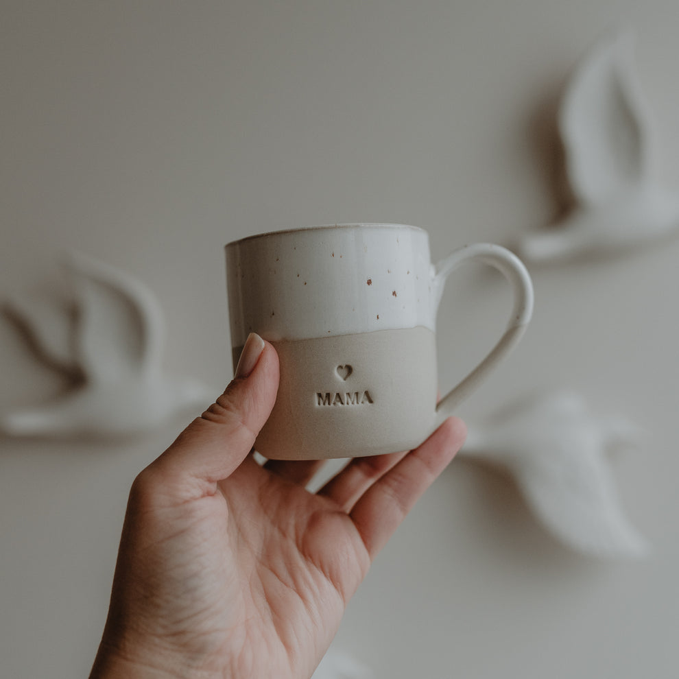 Hand hält eine Cappuccinotasse mit Herzprägung und dem Schriftzug MAMA, vor hellem Hintergrund mit Keramikvögeln an der Wand