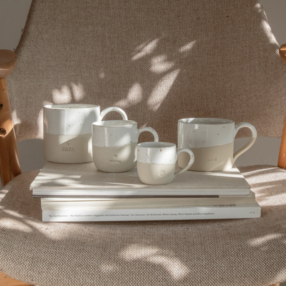 Vier handgefertigte Keramiktassen stehen auf zwei gestapelten Bildbänden auf einem gepolsterten Sessel mit hellbeigem Stoff. Die Tassen sind in natürlichen Beige- und Weißtönen gehalten, oben glasiert mit kleinen braunen Sprenkeln, unten matt. Jede Tasse trägt eine individuelle Prägung: von links nach rechts 'PAPA' mit Herzsymbol, 'MAMA' mit Herzsymbol, eine kleine Tasse mit Smiley-Gesicht und eine größere mit der Prägung 'LOVE'. Sonnenlicht fällt durch Blätter und wirft weiche Schatten auf die Szene