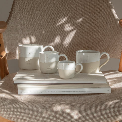 Vier handgefertigte Keramiktassen stehen auf zwei gestapelten Bildbänden auf einem gepolsterten Sessel mit hellbeigem Stoff. Die Tassen sind in natürlichen Beige- und Weißtönen gehalten, oben glasiert mit kleinen braunen Sprenkeln, unten matt. Jede Tasse trägt eine individuelle Prägung: von links nach rechts 'PAPA' mit Herzsymbol, 'MAMA' mit Herzsymbol, eine kleine Tasse mit Smiley-Gesicht und eine größere mit der Prägung 'LOVE'. Sonnenlicht fällt durch Blätter und wirft weiche Schatten auf die Szene