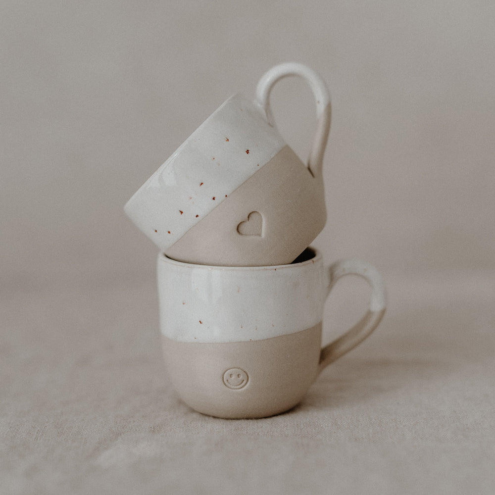 Zwei handgefertigte Keramiktassen stehen auf einem hellen Stoffuntergrund. Eine Tasse mit Herzsymbol ist leicht schräg und ruht auf dem Rand der unteren Tasse, die ein Smiley-Gesicht als Prägung trägt. Beide Tassen haben eine zweifarbige Gestaltung: Der obere Bereich ist glasiert weiß mit braunen Sprenkeln, der untere Teil ist matt und beige.