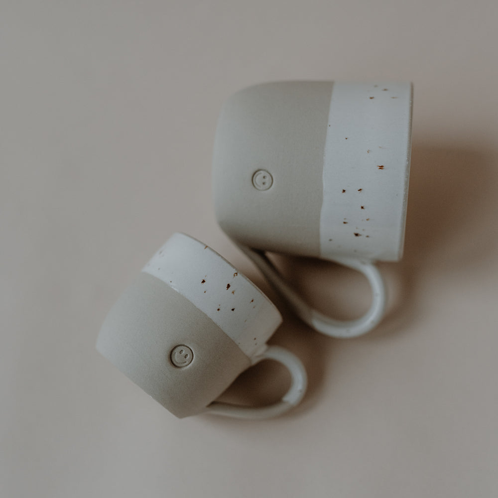 Zwei handgefertigte Keramiktassen liegen nebeneinander auf einer hellen Oberfläche. Beide Tassen bestehen aus naturfarbenem Ton mit einem weißen, gesprenkelten oberen Bereich. Auf der Vorderseite jeder Tasse ist ein kleines, rundes Smiley-Relief eingeprägt. Die Tassen sind unterschiedlich groß – eine größer, eine kleiner – und mit den Henkeln zueinander ausgerichtet.