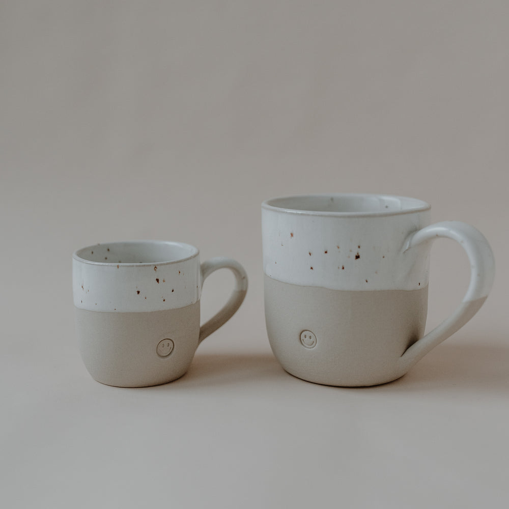 Zwei handgefertigte Keramiktassen stehen nebeneinander auf einer hellen Oberfläche. Die Tassen bestehen aus naturfarbenem Ton mit einem weißen, leicht gesprenkelten oberen Drittel. Auf der Vorderseite ist jeweils ein kleines, rundes Smiley-Gesicht erhaben eingeprägt. Die linke Tasse ist kleiner als die rechte, beide haben einen Henkel auf der rechten Seite.