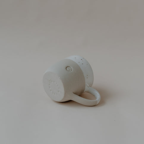 Eine handgefertigte Keramiktasse liegt auf der Seite auf einer hellbeigen Oberfläche. Die Tasse hat einen naturfarbenen unteren Bereich mit matter Oberfläche und einen glasierten, weißen oberen Bereich mit braunen Sprenkeln. Auf der Unterseite ist der Schriftzug des Studios kreisförmig eingeprägt. Auf der Seitenfläche ist ein kleines erhabenes Smiley-Gesicht sichtbar. Der Henkel zeigt nach vorne rechts.