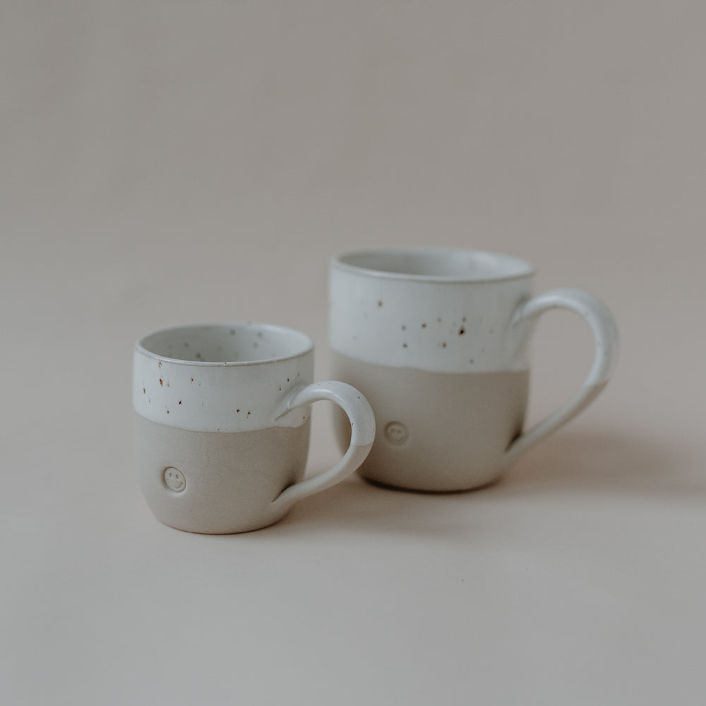 Zwei handgefertigte Keramiktassen stehen auf einer hellbeigen Oberfläche vor neutralem Hintergrund. Beide Tassen bestehen aus unbehandeltem, naturfarbenem Ton im unteren Bereich und einem glasierten, weißen, gesprenkelten oberen Drittel. Auf der Vorderseite jeder Tasse ist ein kleines, rundes Smiley-Symbol erhaben eingeprägt. Die kleinere Tasse steht links, die größere rechts dahinter, jeweils mit dem Henkel auf der rechten Seite.