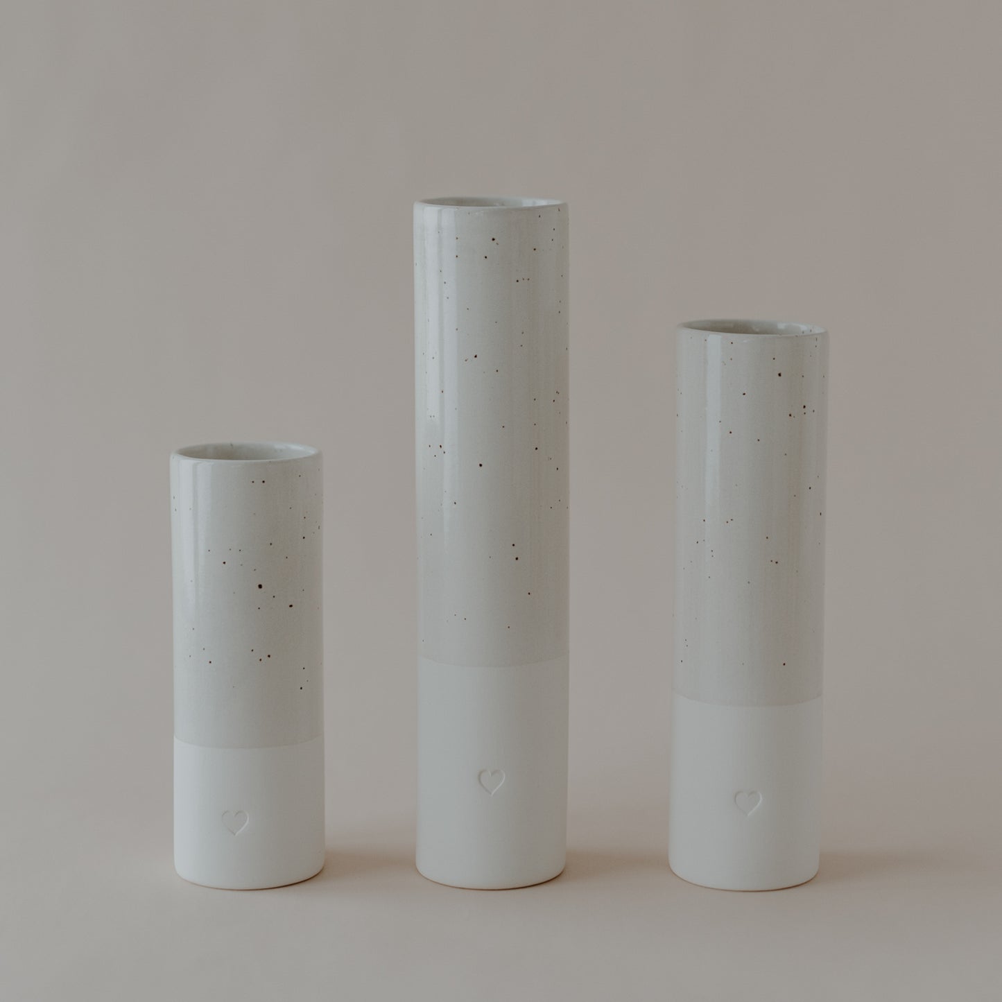 Drei zylindrische Vasen in drei unterschiedlichen Höhen. Im unteren Drittel sind sie weiß mit geprägtem Herz und 2/3 sind lasiert mit dunkelbraunen Sprenkeln.