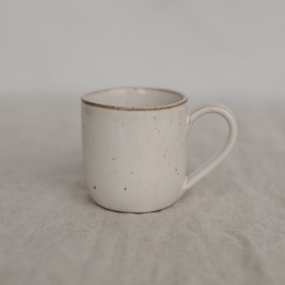 Eine handgefertigte, helle Keramiktasse mit feinen braunen Sprenkeln steht auf einem strukturierten Leinentuch. Die Form ist schlicht zylindrisch mit einem leicht abgerundeten Henkel. Der Hintergrund ist weich und neutral, was der Tasse eine ruhige, natürliche Ausstrahlung verleiht.
