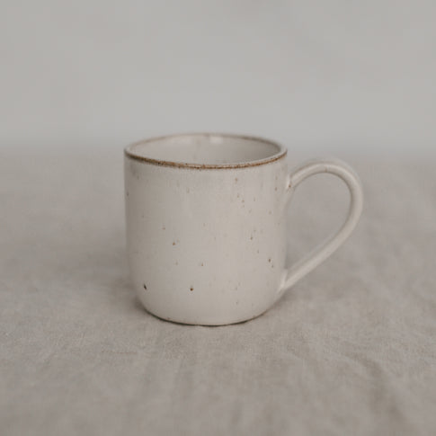 Eine handgefertigte, helle Keramiktasse mit feinen braunen Sprenkeln steht auf einem strukturierten Leinentuch. Die Form ist schlicht zylindrisch mit einem leicht abgerundeten Henkel. Der Hintergrund ist weich und neutral, was der Tasse eine ruhige, natürliche Ausstrahlung verleiht.
