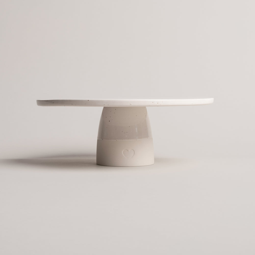 Minimalistische Tortenplatte aus cremefarbener Keramik mit feinen Sprenkeln, auf zylindrischem Fuß stehend; auf der Vorderseite des Fußes ist ein geprägtes Herz sichtbar, vor neutralem Hintergrund fotografiert.