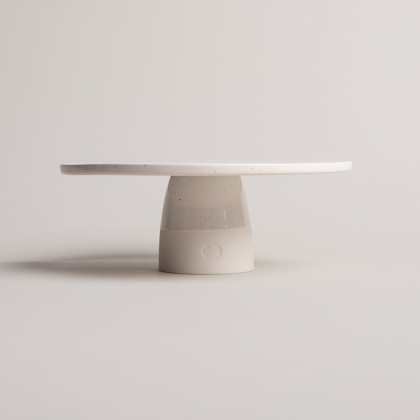 Minimalistische Tortenplatte aus cremefarbener Keramik mit feinen Sprenkeln, auf zylindrischem Fuß stehend; auf der Vorderseite des Fußes ist ein geprägtes Herz sichtbar, vor neutralem Hintergrund fotografiert.