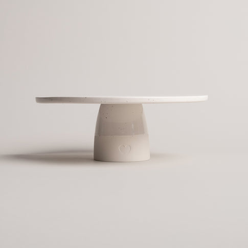 Minimalistische Tortenplatte aus cremefarbener Keramik mit feinen Sprenkeln, auf zylindrischem Fuß stehend; auf der Vorderseite des Fußes ist ein geprägtes Herz sichtbar, vor neutralem Hintergrund fotografiert.