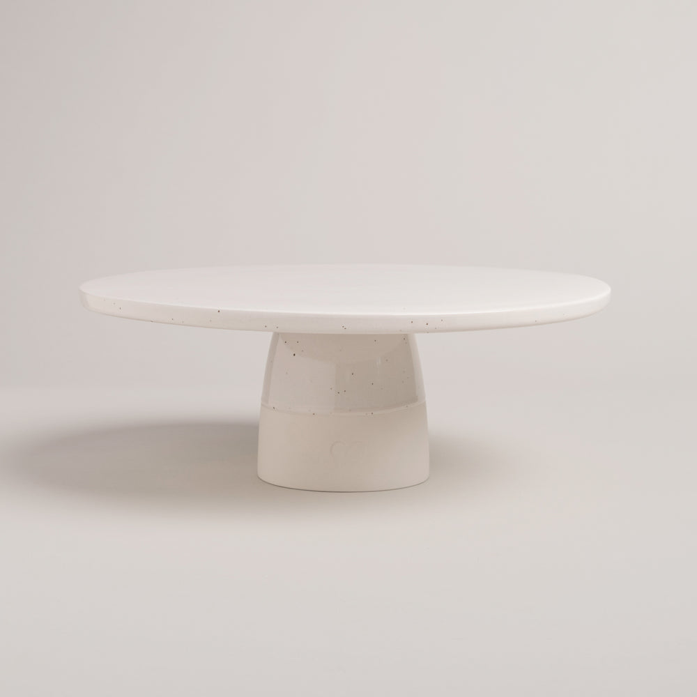 Minimalistische, runde Tortenplatte aus heller, gesprenkelter Keramik mit stabilem Fuß – schlichtes, handgemachtes Design vor hellem Hintergrund.