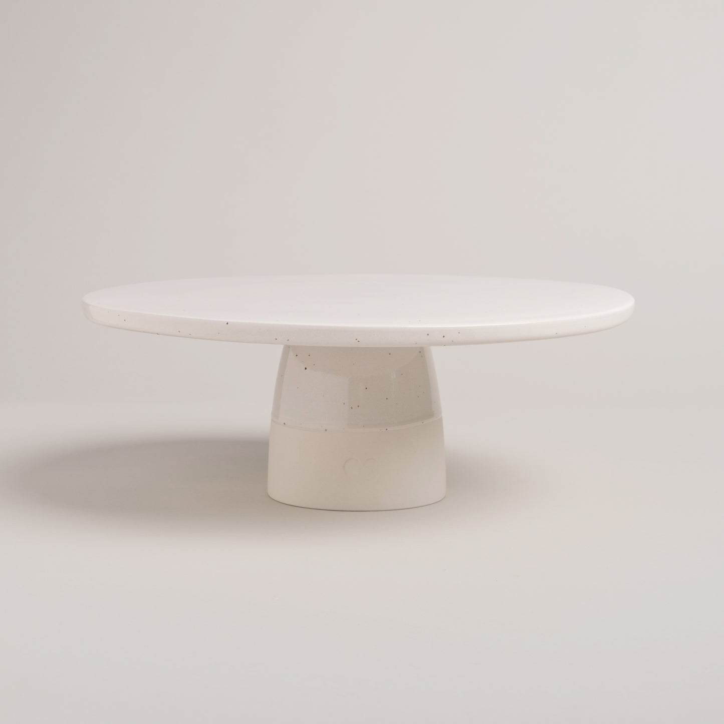 Minimalistische, runde Tortenplatte aus heller, gesprenkelter Keramik mit stabilem Fuß – schlichtes, handgemachtes Design vor hellem Hintergrund.