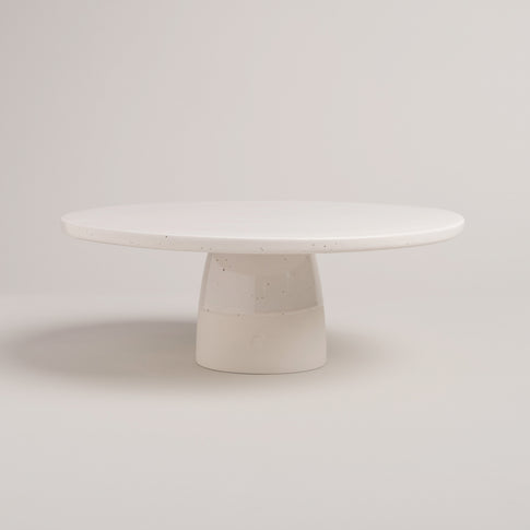 Minimalistische, runde Tortenplatte aus heller, gesprenkelter Keramik mit stabilem Fuß – schlichtes, handgemachtes Design vor hellem Hintergrund.