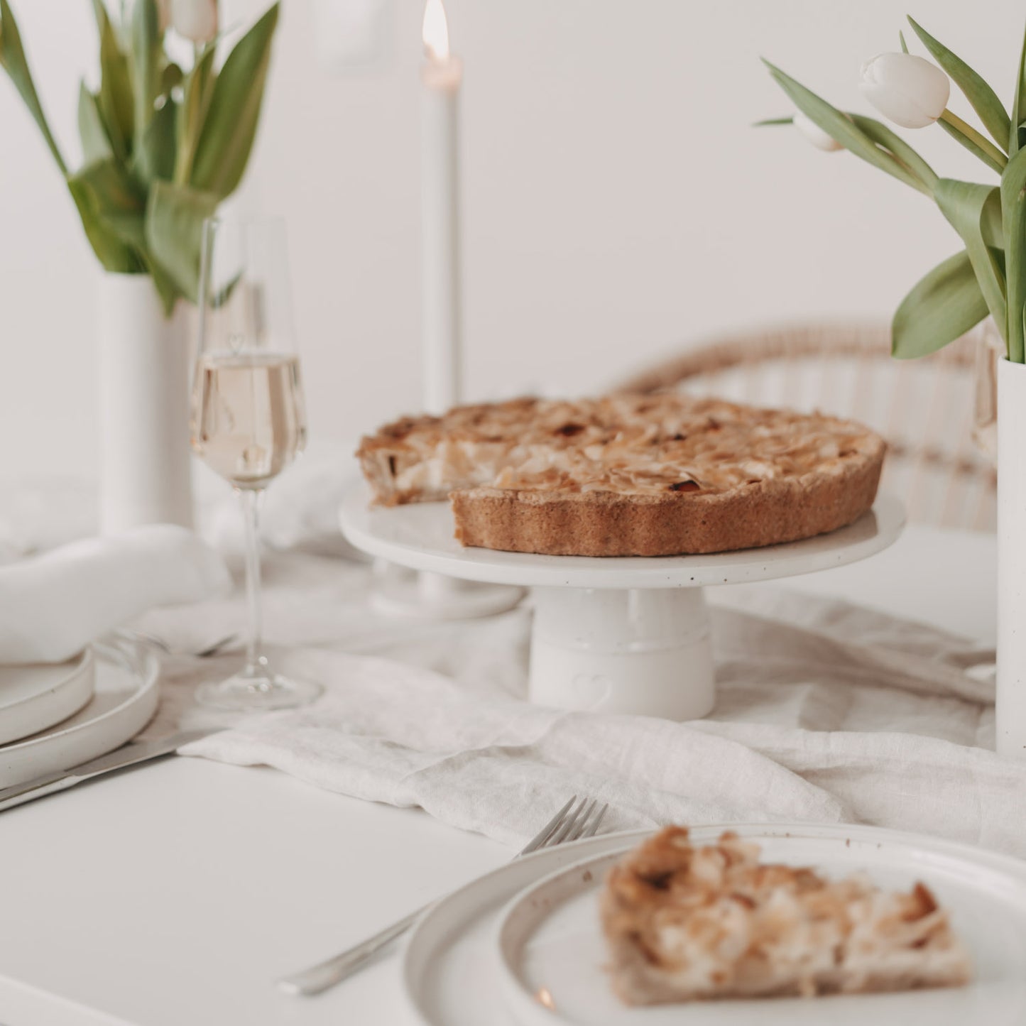 Frühlinghaft gedeckter Tisch mit hellem Leinentuch, Tulpen in weißen Vasen und einer angeschnittenen Tarte auf einer weißen Keramik-Tortenplatte; im Vordergrund ein Teller mit einem Stück Tarte und Besteck, im Hintergrund ein Glas Sekt und brennende Kerzen.