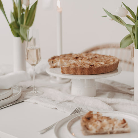 Frühlinghaft gedeckter Tisch mit hellem Leinentuch, Tulpen in weißen Vasen und einer angeschnittenen Tarte auf einer weißen Keramik-Tortenplatte; im Vordergrund ein Teller mit einem Stück Tarte und Besteck, im Hintergrund ein Glas Sekt und brennende Kerzen.