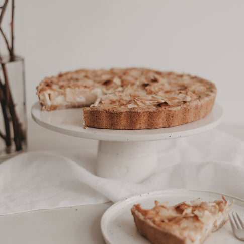 Anschnitt einer goldbraunen Tarte mit Mandelblättchen auf einer minimalistischen weißen Keramik-Tortenplatte mit Herzprägung; im Vordergrund ein Stück Tarte auf einem Teller mit Gabel, im Hintergrund ein weißes Leinentuch und dezente Dekoration.