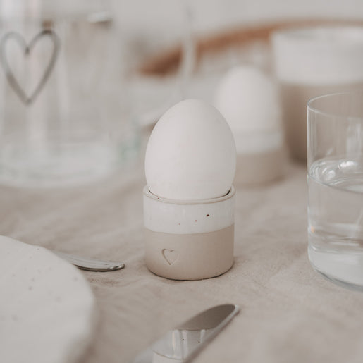 Eierbecher aus handgemachtem Steingut mit Herzprägung und weißem Ei auf gedecktem Frühstückstisch