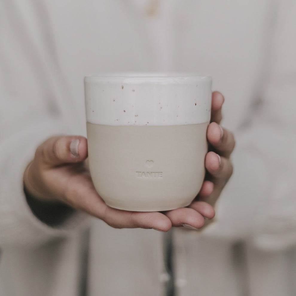 Hand hält einen schlichten, beigen Keramikbecher mit cremeweißer Glasur am oberen Rand. Auf der Vorderseite ist ein kleines Herz und darunter das Wort ‚TANTE‘ eingeprägt. Der Becher wird in beiden Händen vor dem Körper präsentiert – stilvoll, minimalistisch und mit liebevoller Botschaft, ideal als Geschenk für die Tante.