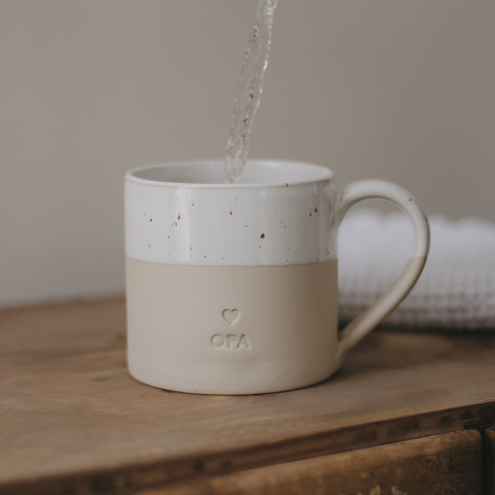 Eine große Tasse aus Keramit mit der Prägung "Opa" steht auf einem Holztisch und es wir Wasser eingegossen.
