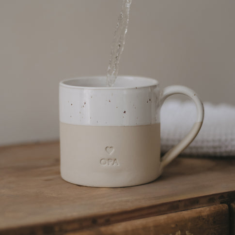 Eine große Tasse aus Keramit mit der Prägung "Opa" steht auf einem Holztisch und es wir Wasser eingegossen.