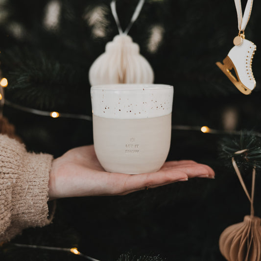 Becher „Let it Snow“ aus Steingut mit Sprenkeln, gehalten vor festlich geschmücktem Weihnachtsbaum.