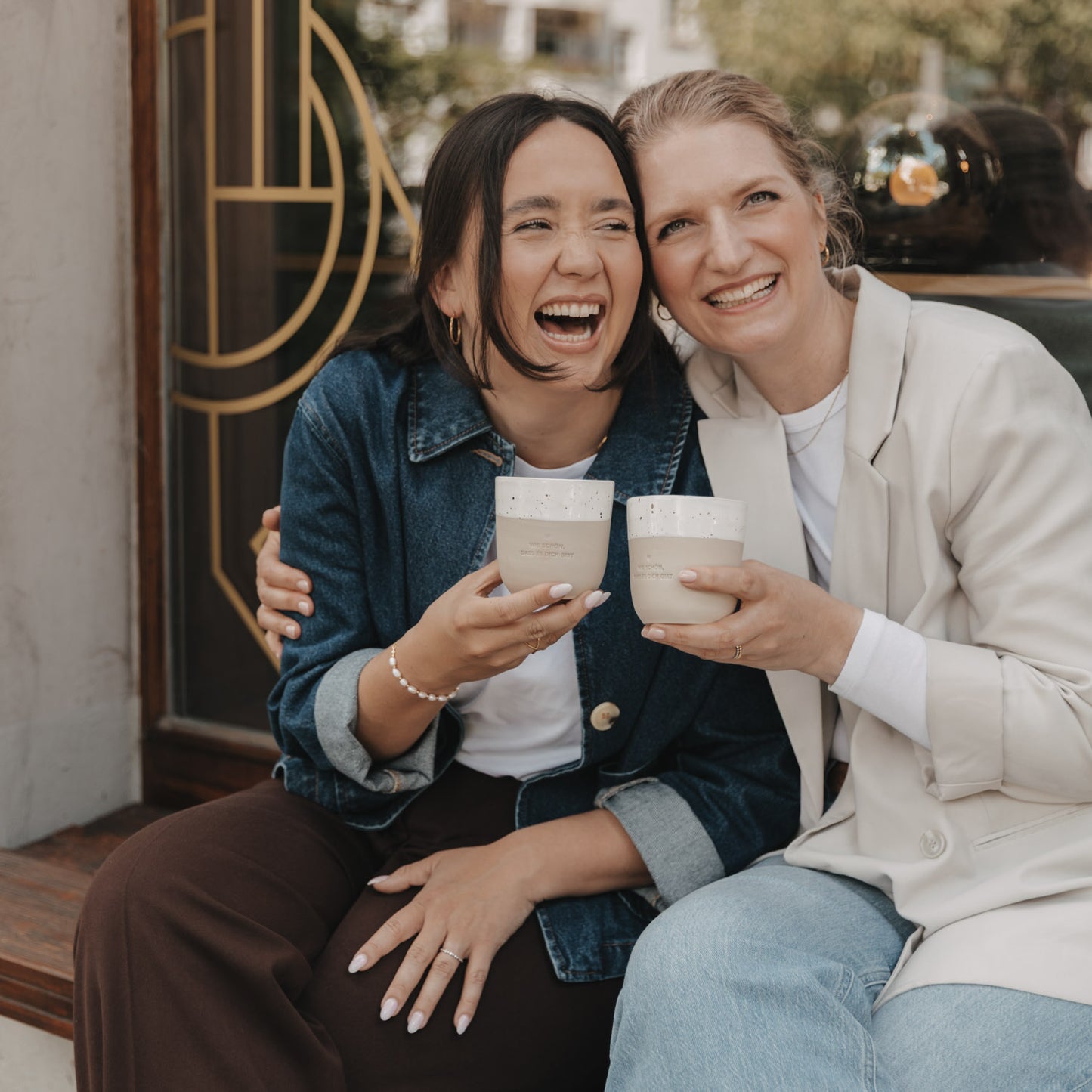 Zwei Freundinnen lachen und freuen sich dicht aneinander gedrückt über die Becher mit denen sie gerade anstoßen. Die Prägung auf beiden Becher lautet "Wie schön, dass es dich gibt" mit einem mittigen Herz darüber.