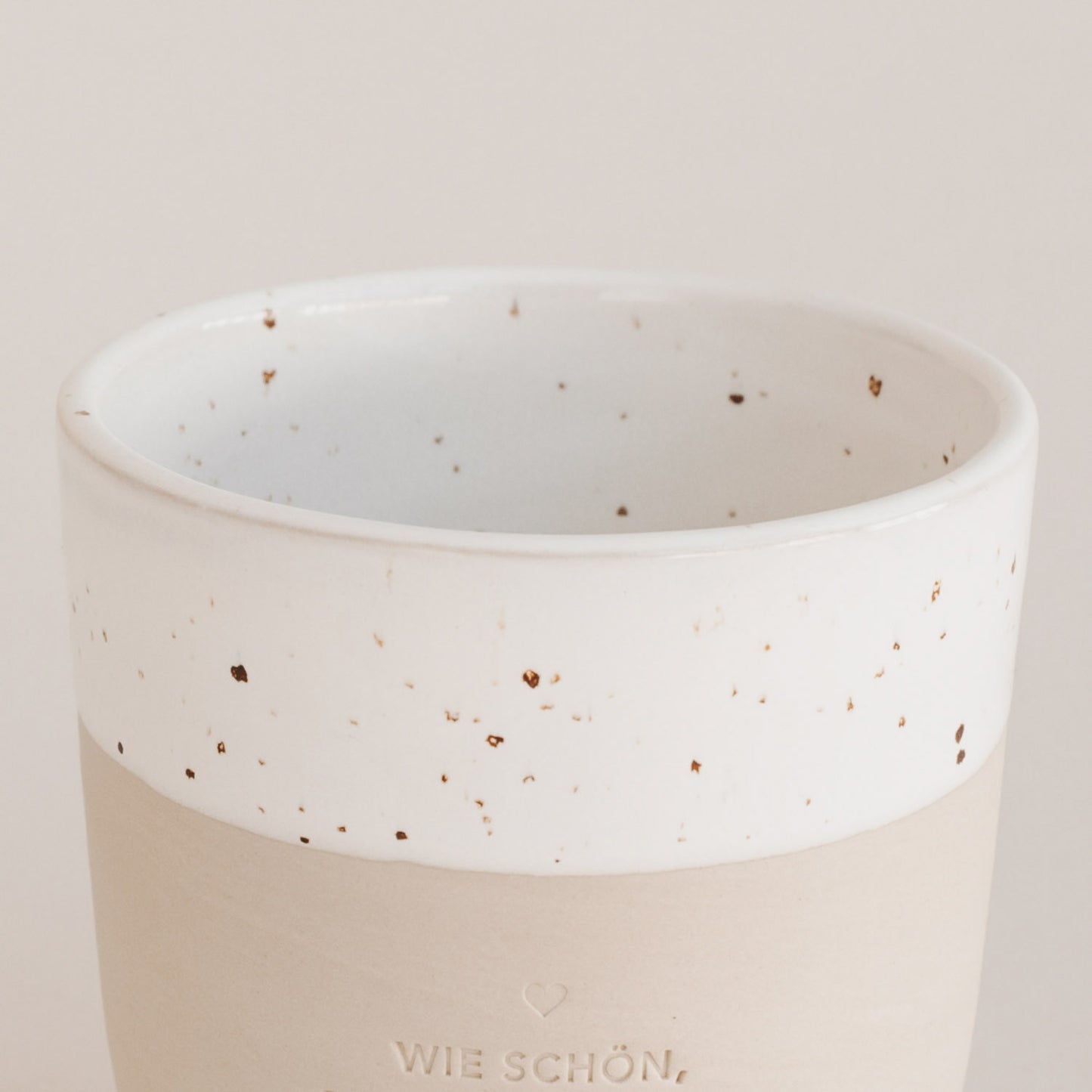 Auf einem grauen Hintergrund steht ein handgetöpfter Becher in Nachaufnahme des oberen Randes, der in weiss mit dunkel braunen Sprenklern lasiert wurde. 