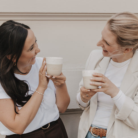 Zwei Freundinnen lachen und freuen sich. Dabei schauen sie sich in die Augen und halten 2 Becher. Die Prägung auf beiden Becher lautet "Wie schön, dass es dich gibt" mit einem mittigen Herz darüber.