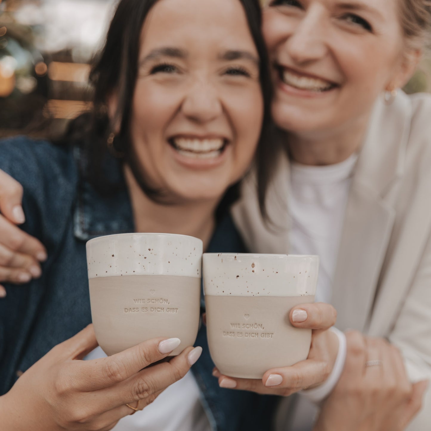 Zwei Freundinnen lachen und freuen sich dicht aneinander gedrückt über die Becher mit denen sie gerade anstoßen. Die Prägung auf beiden Becher lautet "Wie schön, dass es dich gibt" mit einem mittigen Herz darüber.