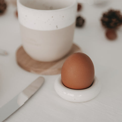 Nahaufnahme: Calma Eierbecher mit braunem Ei neben Becher auf hell gedecktem Tisch mit Besteck