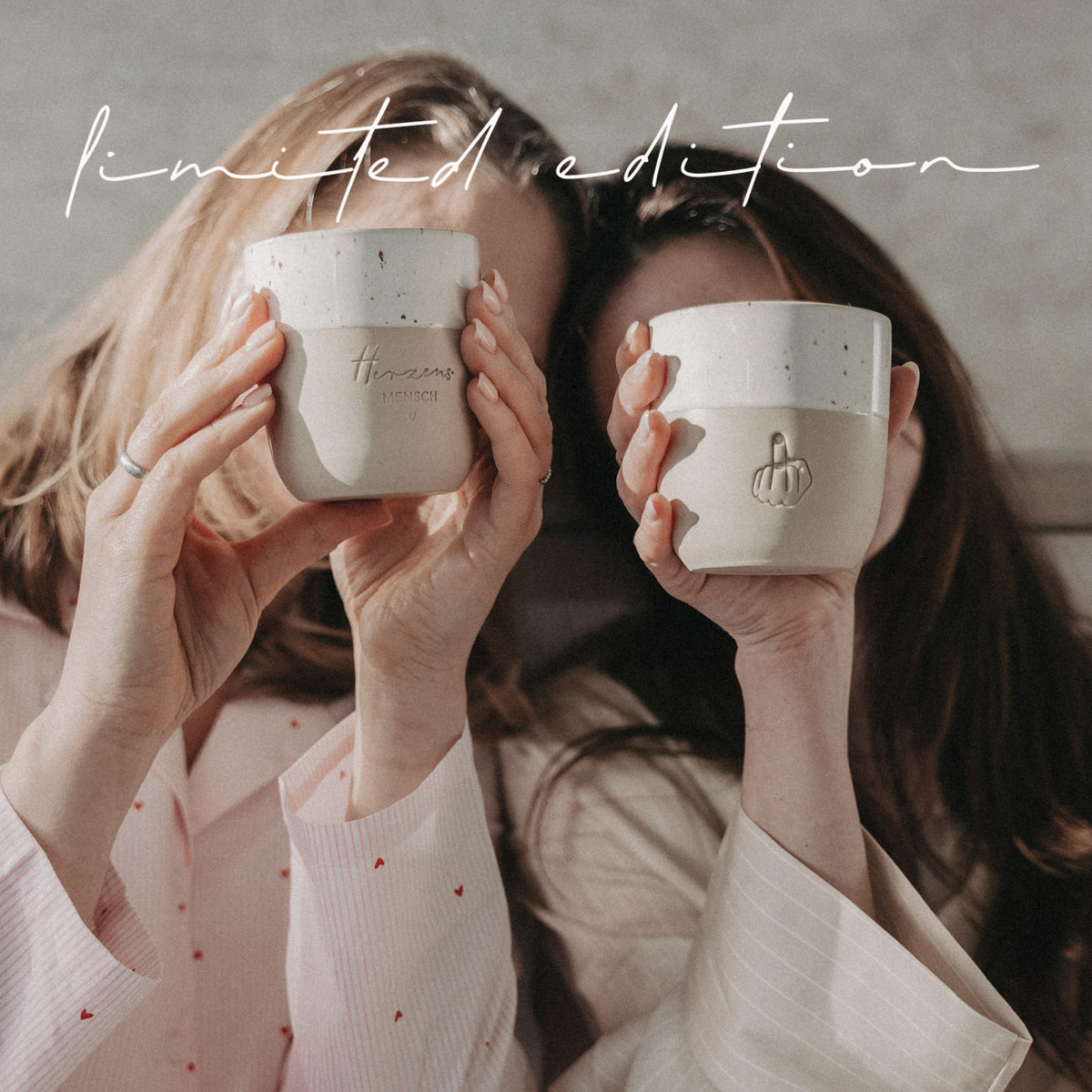 Zwei Frauen halten Herzensmensch- und Mittelfinger-Becher der Limited Edition vor sich, beige-weiß mit Keramikstruktur.