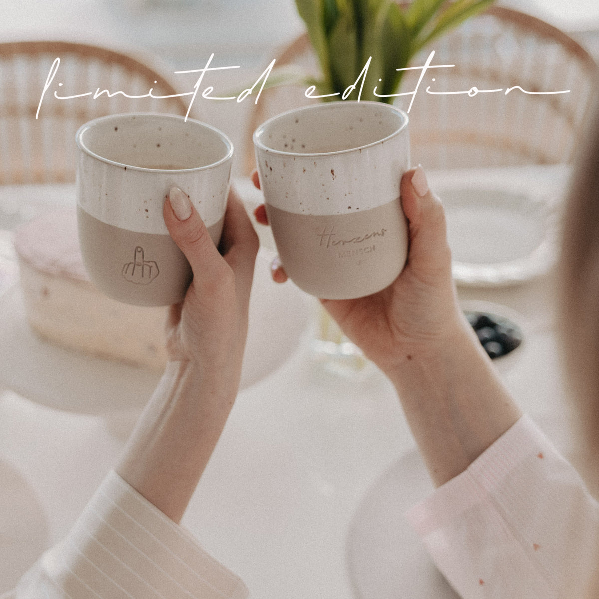 Zwei Hände halten beige-weiße Limited-Edition-Becher mit Herzensmensch- und Mittelfinger-Motiv über einem gedeckten Tisch.