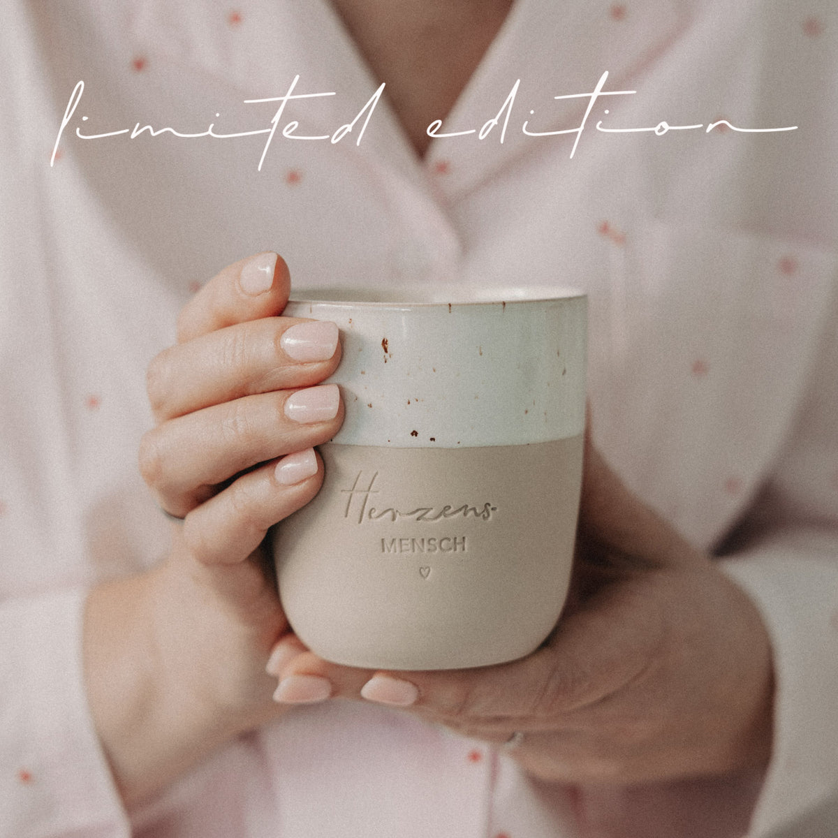 Person hält beige-weißen Limited-Edition-Becher mit Herzensmensch-Gravur nah vor dem Körper in gemütlicher Atmosphäre.